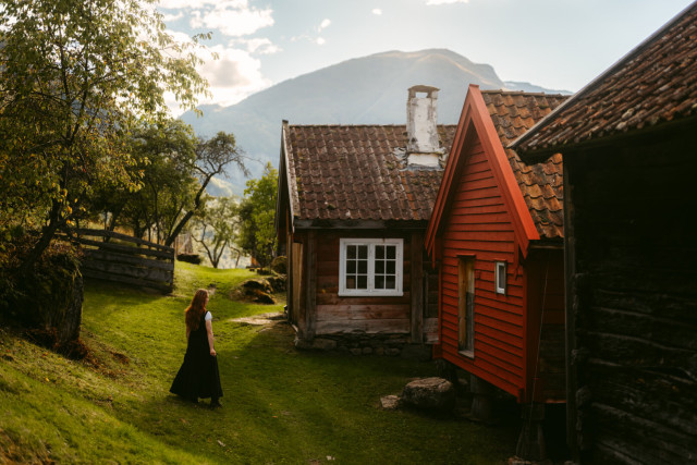The image by European Elopement and Wedding photographer Sturmsucht depicts a tranquil, rustic scene in mountainous Norway, illuminated by warm, golden sunlight. In the foreground, a woman with long reddish-brown hair, wearing a dark flowing dress and a white top, walks up a gentle grassy path to the left, suggesting a peaceful stroll. To the right, two traditional wooden houses stand side by side: the closer one is deep red with horizontal planks and a steeply pitched, moss-covered terracotta roof; the other, slightly behind, is made of weathered brown wood with a prominent white-framed window and a white chimney. A rustic wooden fence winds through trees in the middle ground. In the distance, rolling green hills and a hazy mountain ridge stretch beneath a soft, partly cloudy sky. Lush foliage and scattered trees enhance the natural beauty. The overall atmosphere is one of quiet rural charm, evoking an old village or farmstead nestled in a serene valley.
