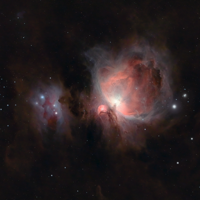 The Great Orion Nebula painted in soft reddish and purple tones opens like a flame to the right of the picture, over a black starry background with brownish dust clouds. On its left, a small irregular opening amidst the clouds reveals a deep dark red region with blue reflections around it known as the running man.