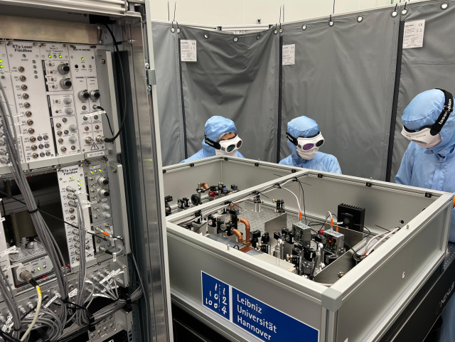 Three persons wearing blue cleanroom clothing and laser goggles stand behind a complicated experimental setup.