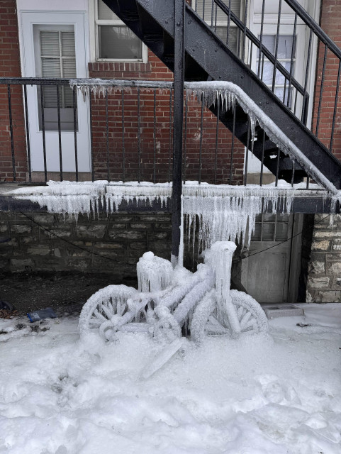 Bicycle leaning against an apartment building fire escape, completely enveloped in ice. Ice on the ground and icicles on the fire escape. Brick and stone building, snowy ground.