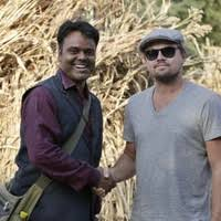 Two people are holding hands in the distance outside against a backdrop of dry, tangled vegetation. The person on the left is wearing a long, dark scarf under a dark waistcoat and carrying a light-coloured shoulder bag, while the person on the right is wearing a grey V-neck T-shirt, a light grey flat-brimmed hat and dark sunglasses. Both are standing close together, with the dry, tall, beige vegetation in the background behind them.