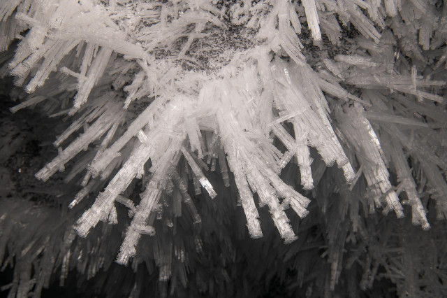 A close-up view of dendritic ice spikes growing downward from the cave ceiling. The crystals are long, branching, and translucent, with sharp edges that catch the light against darker ice in the background.