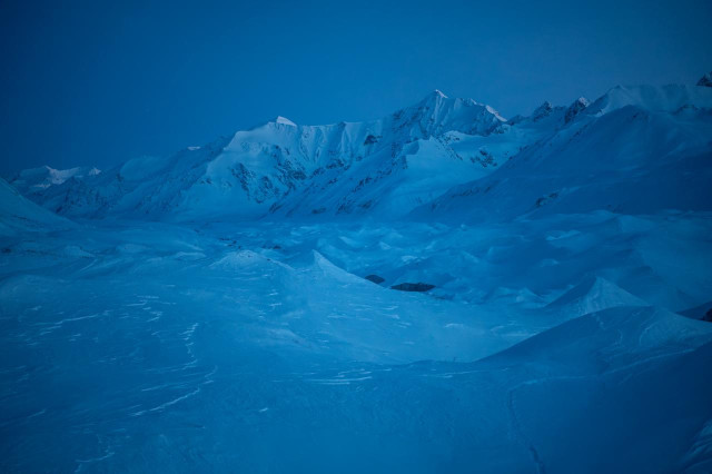 A wide view up the Canwell Glacier valley during blue hour. Snow-covered glacier ice and wind-sculpted snow fill the foreground, while steep, jagged mountains rise in the distance. The entire scene is cast in deep blue twilight, with no direct sunlight remaining on the peaks.