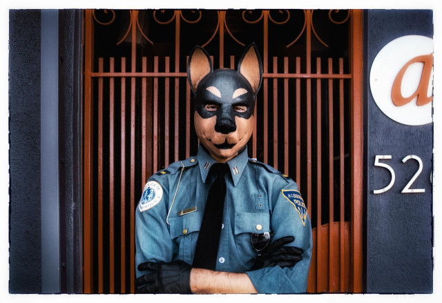 A portrait of a young man in a denim police officer's shirt, black tie, and black gloves. He has his arms crossed and is standing in front of a metal door gate painted the same color as the Golden Gate bridge. 

I should probably also mention he is wearing a full, head covering, rubber Doberman mask. And he has a big smile painted on that mask. His eyes are soft and kind behind that dog mask.