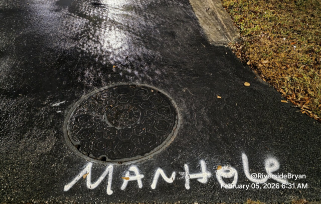 This image is a photograph of a round manhole cover set into dark, wet asphalt or pavement, with prominent white spray-painted text on and around it. 
The primary text, painted in large white capital letters, reads "MANHole" just below the cover. Additional, smaller white text is visible in the bottom right corner, which appears to be a social media handle and a timestamp: "@RiversideBryan February 05, 2026 6:31 AM". 
The manhole cover itself has a raised, geometric hexagonal pattern design, which provides traction for safety and aids in the cooling process during manufacturing. The entire surface of the ground and the cover appears shiny and damp, likely from recent rain. In the upper right portion of the image, there is a small patch of grass and dirt next to a concrete curb. 
The use of spray paint to highlight the manhole could be a form of street art, a protest against municipal neglect (similar to instances where artists use paint to highlight potholes for repair), or a functiona