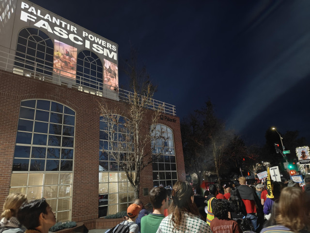 Outside in the evening. An office building with video and the words "Palantir Powers Fascism" projected at top. Below, a crowd of people with signs and candles memorializing people who have died at the hands of ICE.