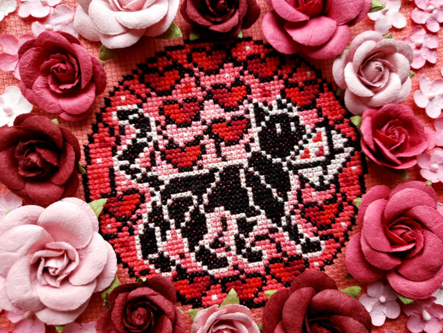 A cross stitch resembling stained glass of a cat carrying a heart-stamped letter with floating hearts around it. The cross stitch is framed by pink and red paper roses.