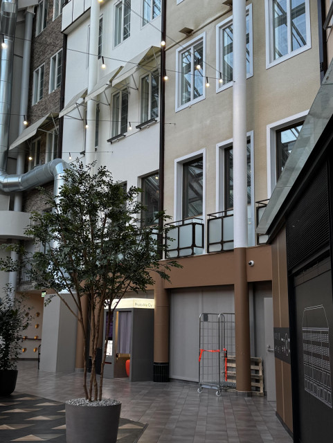 Kolmikulman moderni sisäaula, keskellä iso kasvi/puu ruukussa, beige rakennus ympärillä, riippuvia valoja katossa.