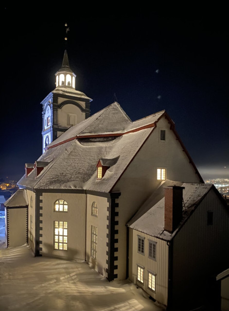 A snowy, illuminated church stands against a dark night sky. Warm light glows from its windows, highlighting steep roofs dusted with snow. A tall tower with a lit belfry rises above the structure, while soft shadows stretch across the snow-covered ground.