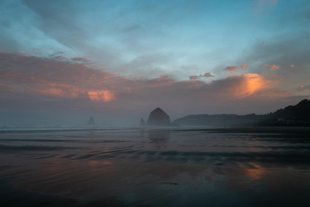 A coastal scene at dawn featuring a beach with wet sand reflecting the sky, characterized by soft hues of blue, pink, and purple. Silhouetted rock formations are visible in the background, along with a distant coastline and gentle waves.