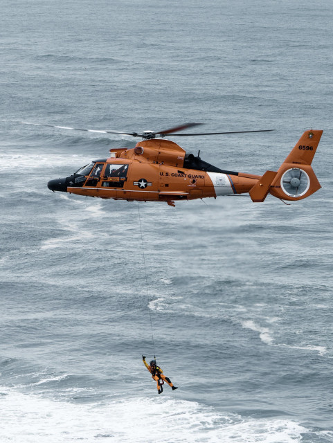 A U.S. Coast Guard helicopter hovers over ocean waves, with a rescuer being lowered via a rope to assist in a rescue mission.