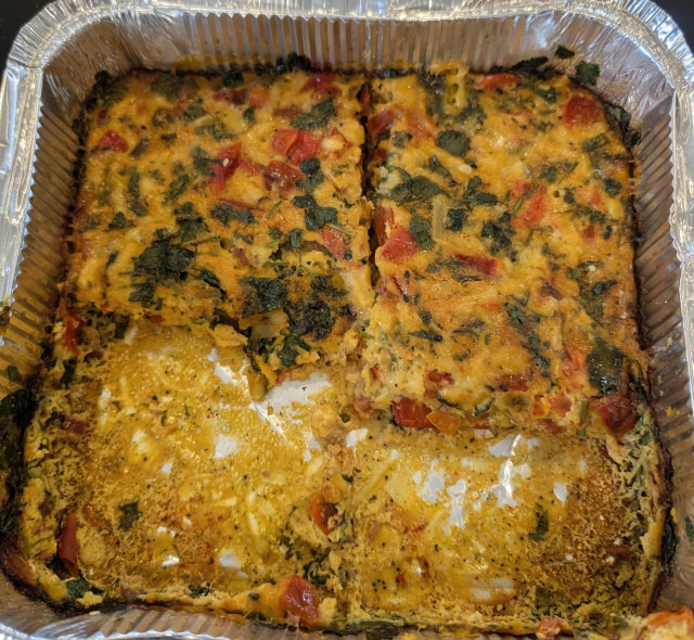 ​A top-down view of a square aluminum foil pan, tightly cropped. The pan contains a golden-yellow egg bake that has been sliced into squares. The top half of the pan holds remaining slices, which are generously speckled with chunks of red bell pepper, cherry tomatoes, and dark green chopped coriander. The bottom half of the pan is empty, revealing the shiny, slightly greasy bottom of the foil container where portions have been served.
