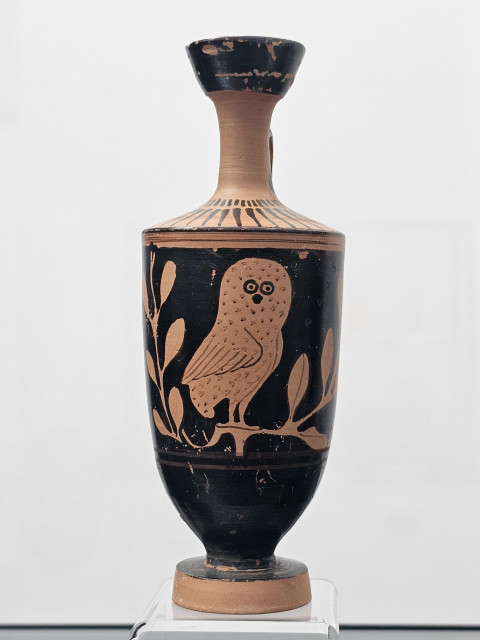 A red-figure lekythos featuring a stylized owl  perched on olive branches on a glossy black body, with reserved red-orange clay for the figure and fine painted details; narrow shouldered form with flaring mouth and footed base, shown on a white stand against a plain background.