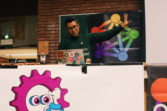 a photo of Maho Pacheco - a man with dark hair, a beard and glasses - standing behind the stage at FOSDEM, looking at a computer screen, smiling and with his left arm stretched out