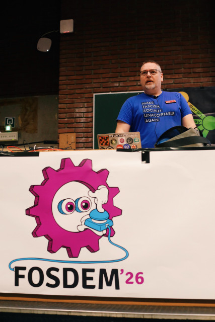 a photo of James Smith standing on stage at FOSDEM. He's wearing an amazing blue t-shirt that reads "make fascism socially unacceptable again"
