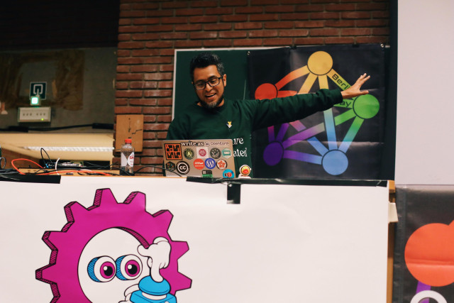 a photo of Maho Pacheco on stage at FOSDEM. He's looking down at a computer screen, smiling, with his arm stretched out towards the screen