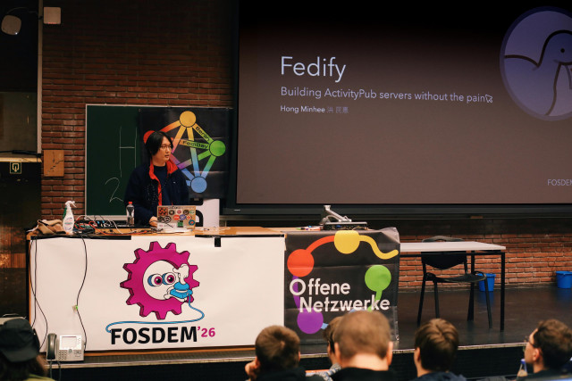 a wide shot of Hong Minhee standing on stage at FOSDEM with the screen reading "Fedify - Building ActivityPub servers without the pain"