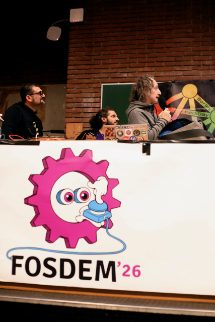 a photo of Michele Agostinelli, Fra and Filippo Della Bianca on stage at FOSDEM during their talk... they're looking off to the right where the big screen is