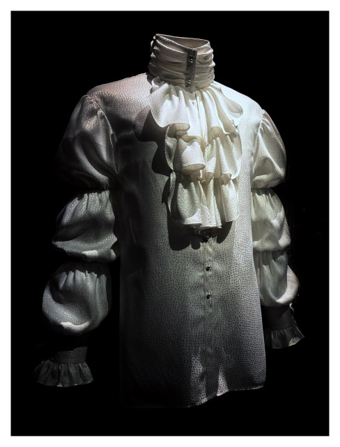 Prince’s white silk shirt from his 1980s Purple Rain era, with puffed sleeves and a high collar with a fall of ruffles, on display in a museum exhibit against a black backdrop. Photo taken at the recent exhibition “Superfine: Tailoring Black Style” at the Metropolitan Museum of Art in NYC.