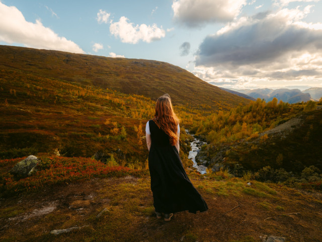 This image by European Elopement and Wedding Photographer Sturmsucht captures a female person standing with their back to the viewer, adorned with long, reddish-brown hair and a dark dress, as they gaze upon a sprawling autumnal Norwegian landscape. The foreground and rolling hills are a rich tapestry of fall colors, featuring golden-brown grasses, deep red and orange shrubs, and clusters of vibrant yellow-leafed trees. A winding river cuts through the mid-ground, its waters appearing bright against the colorful terrain. The sky above is a dynamic mix of blue patches and dramatic white and gray clouds, with sunlight casting a warm, golden glow across the scene, creating a serene and expansive wilderness atmosphere.