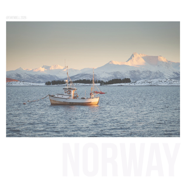 A very small fishing boat is moored in a fjord with distant mountains in the background catching a little pink light at the end of the day