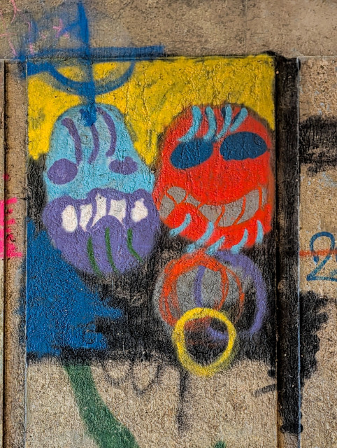 A piece of expressive, colorful graffiti painted onto a rough, textured concrete surface. The style is raw and illustrative, featuring two stylized, monster-like heads.

On the left is a blue and purple figure with a wide, toothy grin and dark circular eyes. To its right is a vibrant red figure with large blue oval eyes and a similar wide-mouthed expression. Both characters have blue, hair-like streaks extending from the tops of their heads.
 
The characters are set against a bright yellow and blue background, which is framed by thick, dark vertical lines. This creates a "panel" effect, making the artwork stand out from the surrounding gray concrete.
