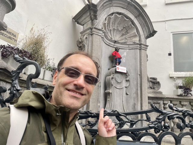 A person smiles in the foreground, pointing at a small figure dressed in a red outfit, positioned on a decorative wall. Nearby, there is a sign indicating "Camera Security." The background features ornate architectural details and some greenery.