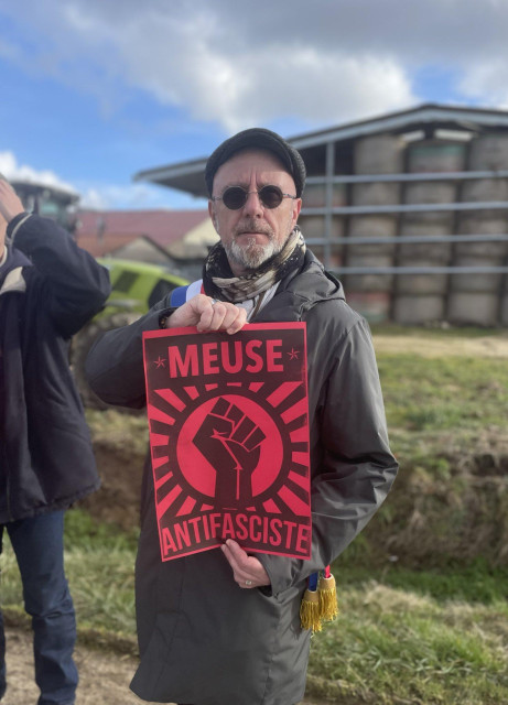 A man in a black coat, with a white beard and sunglasses, holds a red placard with the raised fist and text reading MEUSE ANTIFASCISTE