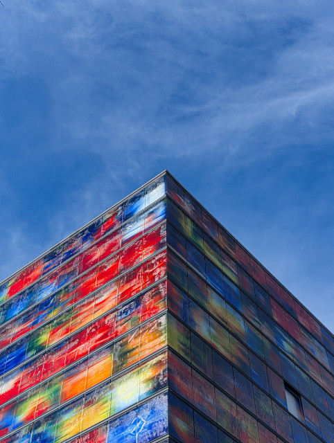 A low-angle shot of a modern building facade, characterized by its vibrant, multicolored glass panels. The architecture is sharp and geometric, dominated by the 90-degree corner of the structure that points toward the top of the frame.

The building (the Netherlands Institute for Sound and Vision) is covered in a grid of glass panels. Each panel features abstract, painterly streaks of color, primarily saturated reds, deep blues, bright yellows, and greens. The glass panels feature iconic images from Dutch television history, blurred into this colorful abstraction.