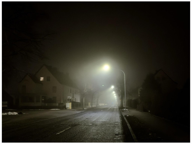 A foggy street scene at night with dimly lit street lamps illuminating a deserted road. Houses are visible in the background, partially obscured by the fog.