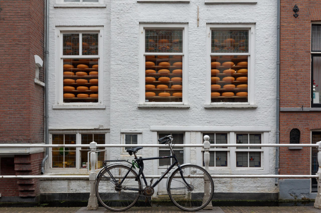 Amsterdam: a white building with large windows displays numerous round orange cheeses. In the foreground, a black bicycle is parked beside a white railing. The scene features brick walls on either side, creating an urban atmosphere.