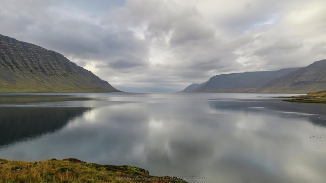A colour photo of a fjord with mountains on either side. The foreground is the water's edge, a rocky outcrop with autumnal grasses over it. The water is ruffled by a breeze, but is reflecting the mountains. There are a few birds on the surface on the right. The peaks level out at the mouth of the fjord, coming down to meet the water. The sky is grey and overcast but with a few thinner patches, so that clear sky can almost be seen.