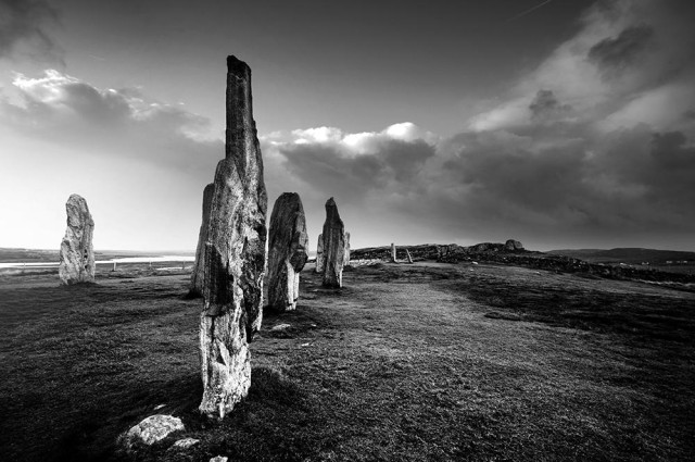 Atmospheric black and white photo of tall standing stones at sunset, with dark clouds overhead.