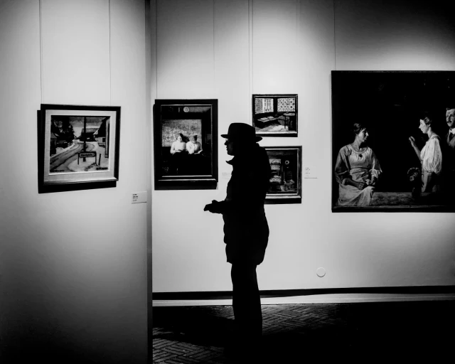 Die Silhouette einer Frau mit Hut steht vor den Gemälden verschiedener Künstlerinnen im Museumsberg Flensburg. 
.
The silhouette of a woman wearing a hat stands in front of paintings by various female artists at the Museumsberg Flensburg. 
.