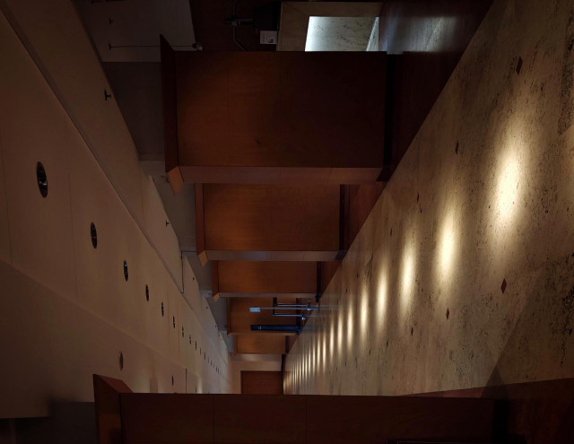 A tilted photo of a hotel corridor with warm-toned wooden panels. The floor is stone with evenly spaced light spots. 
