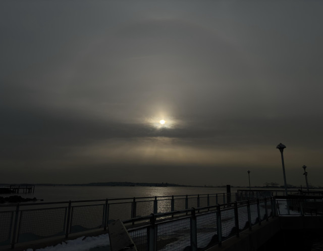 A view from a pier overlooking the waters of a wide bay. A halo of pale light arcs high over the setting sun as it shines over a strip of dark clouds. Below, golden crepuscular rays fan out in a luminous band before being obscured by more gray clouds at the horizon. Directly beneath the sun, a column of sunlight sparkles on the water. In the foreground are guardrails flanking the pier’s snow-covered walkway as it recedes to some lampposts at right frame.