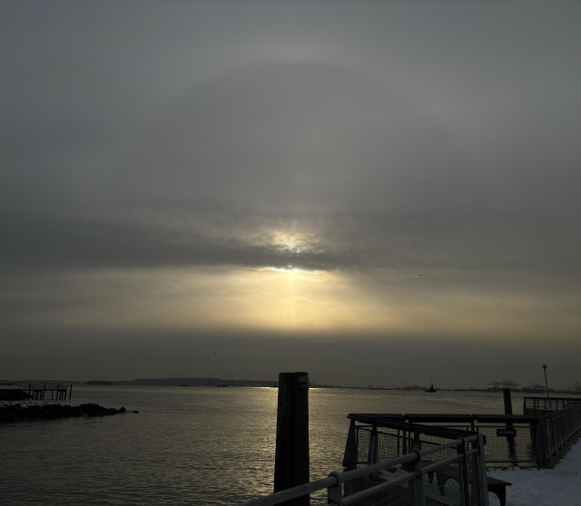 Another view of the sunbow over the pier. The strip of gray clouds obscures the sun itself, without diminishing the halo above or the crepuscular rays below — a strikingly dramatic effect. Directly below the sun is the silhouette of a wooden pillar rising out of the bay, surrounded by the glow of the sun sparkles on the water.