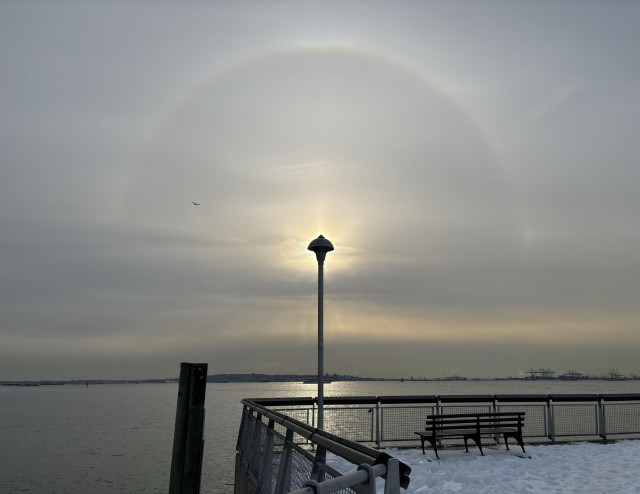 A third view of the sunbow over the pier, with the sun hidden behind the head of a lamppost as the halo arcs overhead. Sunlight glows around the lamp head as the lamppost’s lower half is framed by sun sparkles on the water. To the left is a wooden pillar in the bay; to the right is the pier’s snow-covered walkway, with a bench facing the water and the far shore.