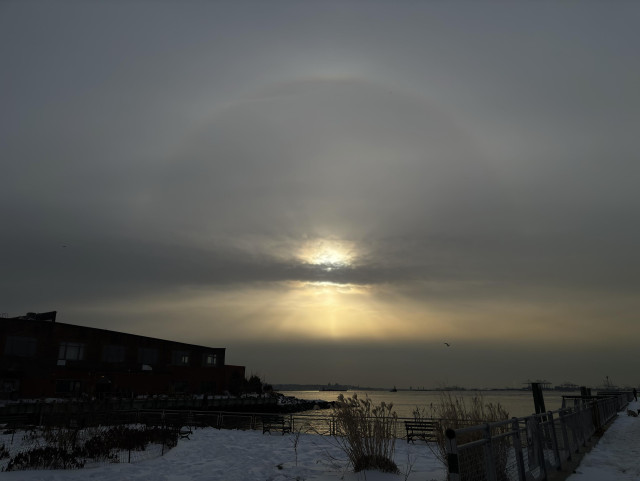 Last in a series of views of a sunbow over a wide bay. A strip of clouds obscures the sun without dimming the arc of the halo above or the crepuscular rays below. In the foreground is a section of a park, with clumps of plants poking out of the snow-crusted ground. At left and right respectively, a low warehouse and a snow-covered pier push out into the water.