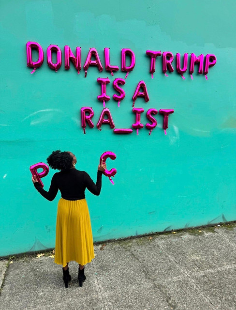 Young black woman holding a letter shaped balloon in each hand. One is a P an one is a C. She’s looking up at a letter balloon message on a wall that reads “Donald Trump is a ra_ist.”