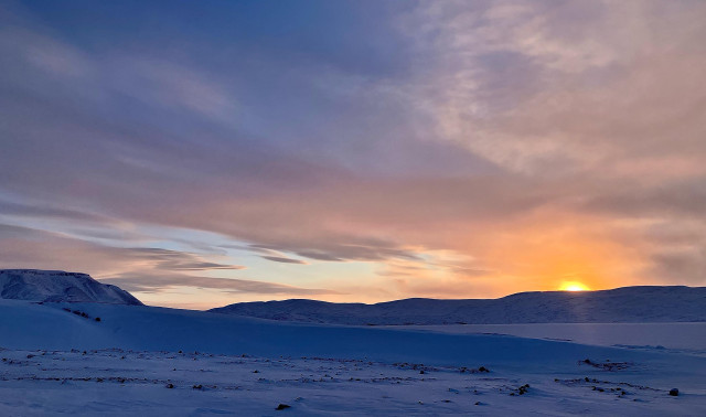 The sun peeks over hills across the frozen ocean. Clouds across the sky are orange, pink, mauve and grey. 