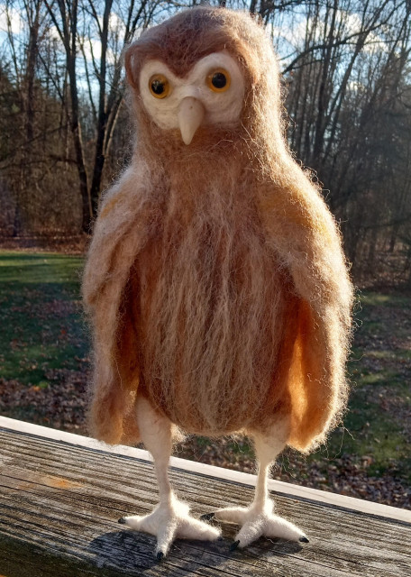 A felted owl sculpture standing on the rail of a deck outdoors. It is tan, brown and white. 