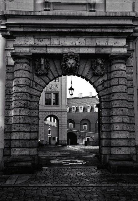 A striking black-and-white photograph of the Stallhof in Dresden, framed by a grand stone archway. The archway, adorned with intricate carvings and decorative elements, leads into a historic courtyard. The cobblestone path under the archway draws the eye towards the buildings beyond, which feature classic architectural details such as arched windows and tiled roofs.