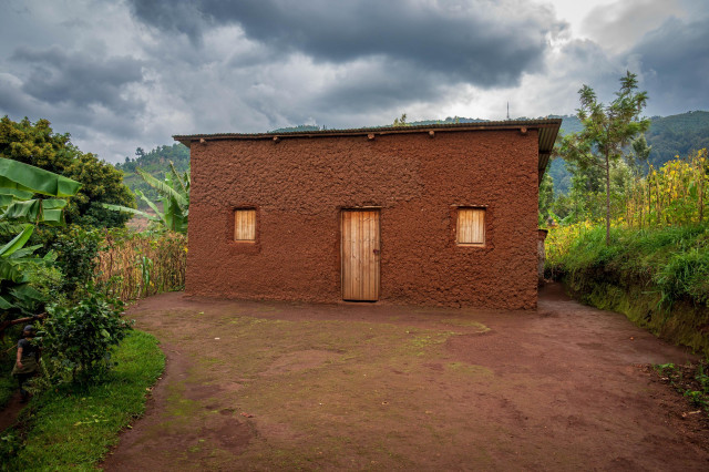 A single-story mud house with a wooden door and two small windows, surrounded by greenery and a dirt path. The sky is overcast with clouds, and hills are visible in the background.