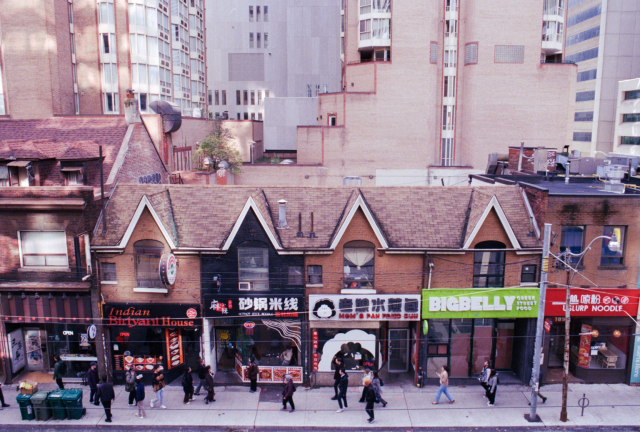 People walk by the shops on Dundas Street in downtown Toronto. While the shops are in older two and three story buildings, massive condo and office towers rise behind them. 