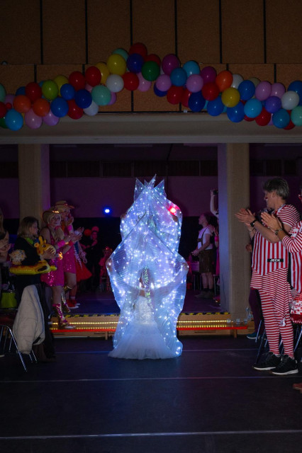 Eine Reihe von Frauen in weissen Kostümen, die mit Lichterketten durchzogen sind, positionieren sich um auf die Bühne zu gegen. Die vordere kniet, da hinter stehen die anderen.
rechts und links ist Publikum zu sehen, oben am Hallendach bunte Ballons zur Dekoration. 