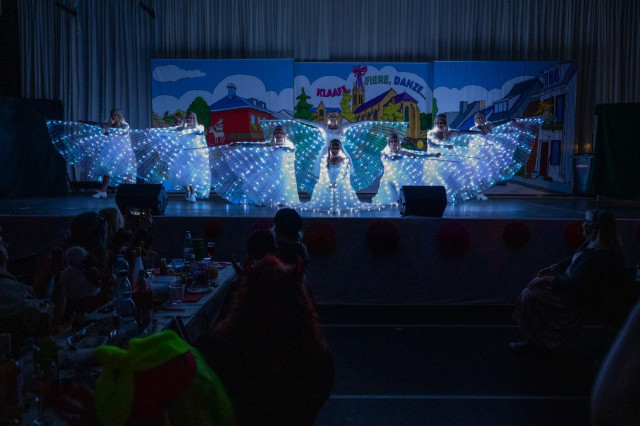 Die Schlussformation eines Tanzes, die Gruppe hat sich auf der Bühne aufgestellt. Die vordere kniet, während die anderen rechts und links ihre Arme in eine Richtung, die hintere hat beide Arme ausgestreckt. Im Hintergrund ist eine Bühnendekoration zu sehen. Rechts und links unten, Teile des Saalpublikums. 