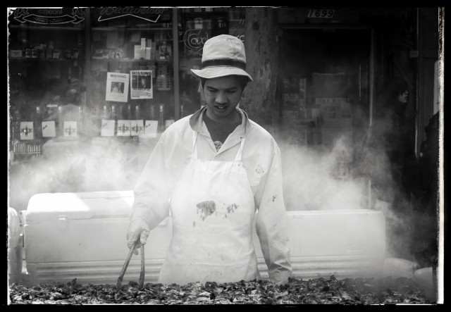 A monochrome photo of young man on a Haight Street sidewalk, with an apron, cap, and tongs in his hand. He is standing over a smokey grill full of skewered chicken. The smoke forms a cloud around him (and the smell of teriyaki bbq fills the air). Behind the man, you can see a storefront window with shelves of food and a couple of posters of missing children. The Haight Ashbury neighborhood, hippy central, is known for runaways.
