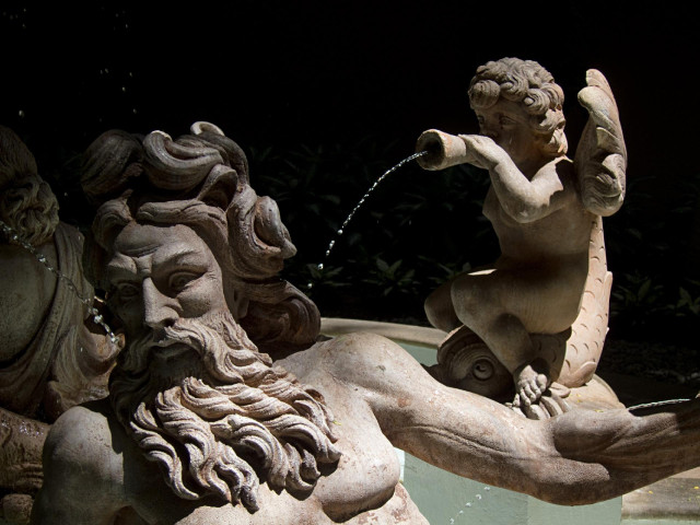 A photo of a stone fountain with sculptures depicting human, probably mythological, figures. White stone and some water streams are shown against a very dark background.
