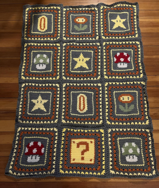 A crochet blanket with pixel images from Mario: a question mark block, red mushroom, green mushroom, fire flower, coin and star.